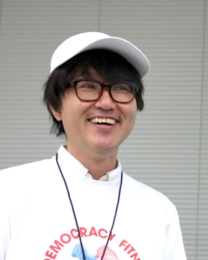 森本康仁。白いキャップと眼鏡、DEMOCRACY FITNESSのTシャツで笑っている